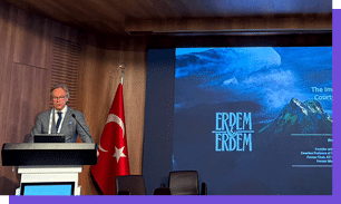 Pessoa adulta vestindo terno e gravata est em um p lpito falando em um ambiente interno com parede de madeira e uma bandeira vermelha com estrela branca ao lado, enquanto uma tela exibe o texto “ERDEM & ERDEM" e uma imagem de montanhas ao fundo.  Yapay zeka taraf ndan olu turulan a  klama